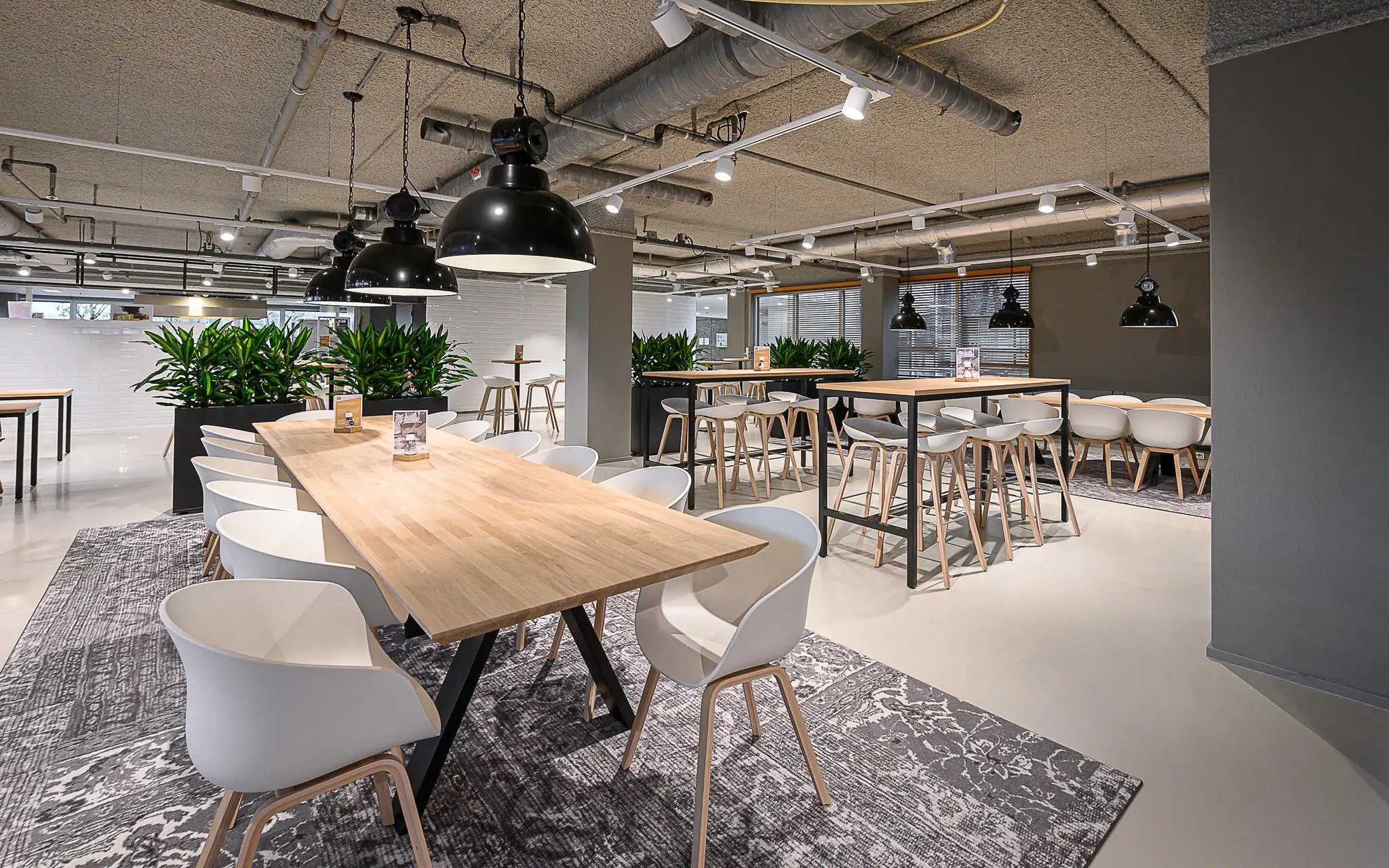 Moderne, open kantoorkantine met houten tafels, witte stoelen, hanglampen en groene planten bij Rivium Quadrant.